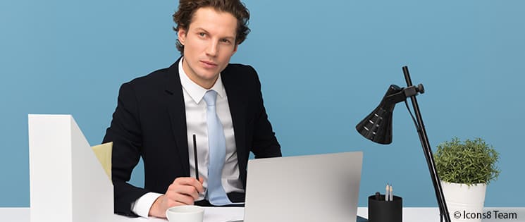 Un homme en costume de bureau est assis à son bureau. Devant lui, un ordinateur portable, un classeur, une lampe de bureau, un pot de crayon, une tasse à café et un pot de plante verte.