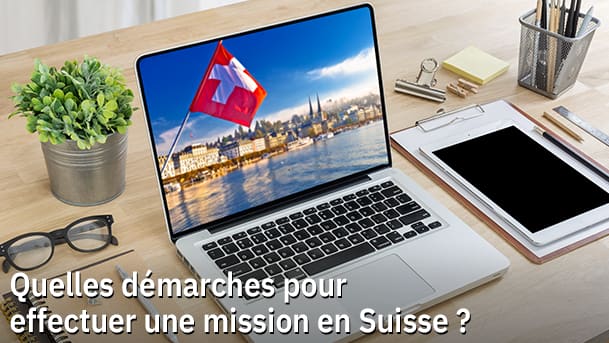 Un ordinateur portable est posé sur un bureau en bois. À l'écran est affichée une image d'un paysage, une ville au bord d'un lac, et un drapeau Suisse en avant plan. Le sujet est "Article sur "Quelles démarches pour effectuer une mission en Suisse ?"